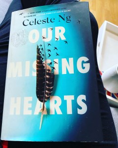 The front cover of the book "Our Missing Hearts" showing the title, the author name. The cover is blue with a bird's feather on the front. The feather is breaking down from the top, with the fibres of the feather turning into little birds and flying away. 