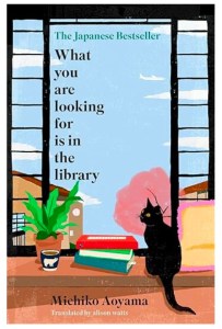 This is the front cover for the book What You Are Looking For Is In The Library by Michiko Aoyama. The cover illustration shows a window over looking houses and a cherry tree. A black cat sits on a desk or shelf in front of the window next to a pile of books and a pot plant.