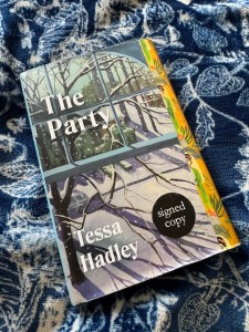 This image shows the front cover of the book The Party by Tessa Hadley. The cover depicts a window with highly decorated yellow curtains on its right hand side. The decorations on the curtains are of a dark haired woman dressed in a green robe with red flowers in her hair. There are leaf drawings and other showers, and square shapes on the curtains too.

Out of the window the view is of a wintry street scene. You can just about see the houses opposite, in front of them are hedges and, dominating the view from the window is a bare, snowy tree, its branches reaching towards the window.