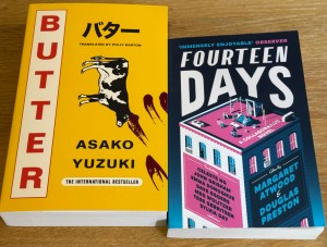 2 books sit side by side on a wooden table. One book, Butter by Asako Yuzuki is a yellow book. It has the book's title on it going down the left side of the book in red capital letters, and the rest of the writing including Japanese characters in black text. There is a cow on the front cover as well as some smears of something - could be blood?
Fourteen Days is the book on the right hand side. It is a book with text and illustrations in blues, whites and pinks. The illustration is of a square apartment block, with people sitting on the top of the roof. The names of the authors of the book are placed on the building in what appear to be billboard adverts.